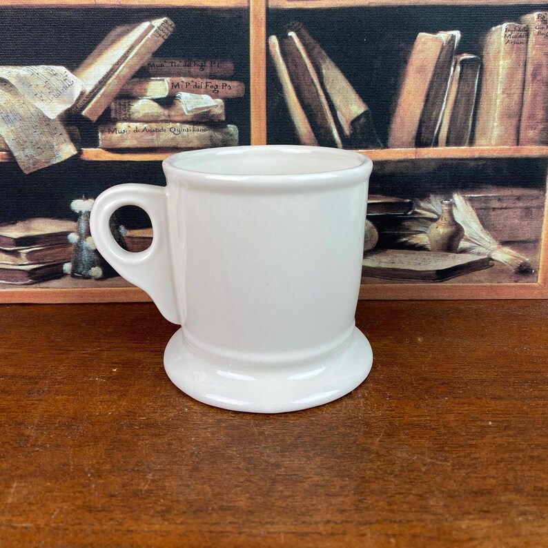 May include: A white ceramic shaving mug with a handle. The mug has a wide base and a rounded top. The handle is a loop shape. The mug is clean and simple, with a classic design.