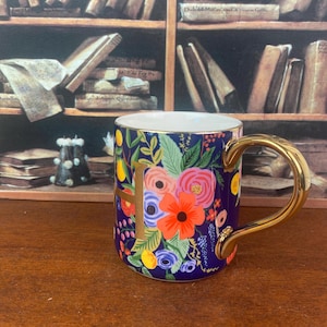 May include: A ceramic mug with a gold handle and the letter "H" in gold. The mug features a dark blue background with a floral design in shades of pink, orange, yellow, and green. The mug is sitting on a wooden surface.
