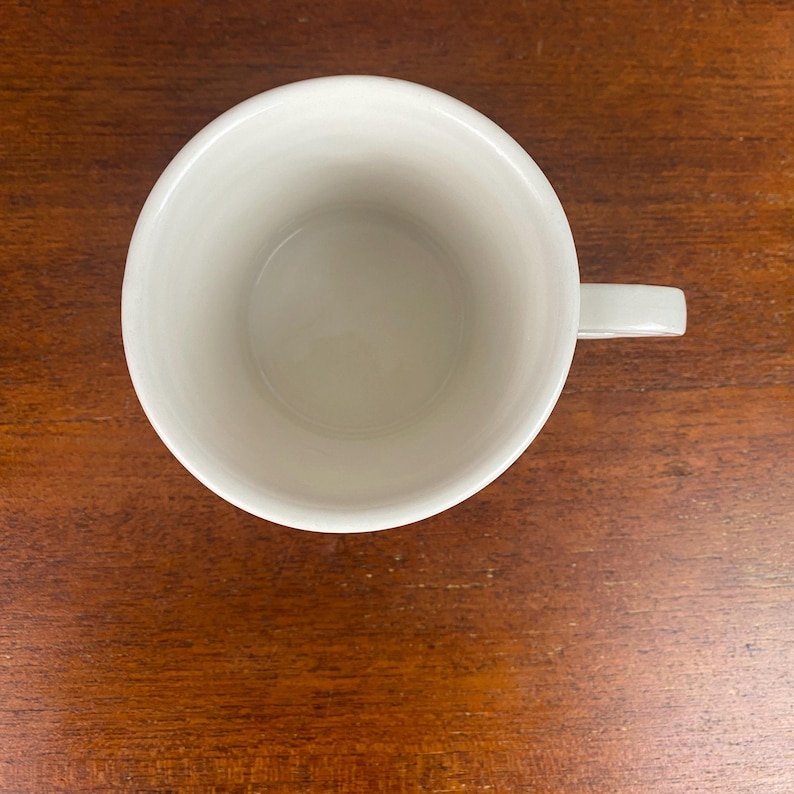 May include: A white ceramic mug with a handle, viewed from above. The mug is empty and placed on a dark brown wooden surface. The interior of the mug is smooth, and the handle is a simple, curved design.