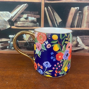 May include: A ceramic mug with a gold handle and rim, featuring a vibrant floral design on a navy blue background. The flowers are in shades of pink, orange, yellow, and blue. The mug is likely used for beverages.