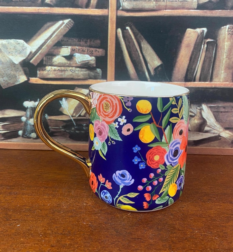 May include: A ceramic mug with a gold handle and rim, featuring a vibrant floral design on a navy blue background. The flowers are in shades of pink, orange, yellow, and blue. The mug is likely used for beverages.
