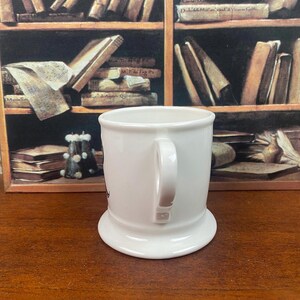 K&ouml;nnte beinhalten: Eine wei&szlig;e Keramiktasse mit rundem Boden und gebogenem Henkel. Die Tasse hat ein schlichtes, elegantes Design, geeignet f&uuml;r Kaffee oder Tee. Der Hintergrund zeigt ein Holzregal mit B&uuml;chern.