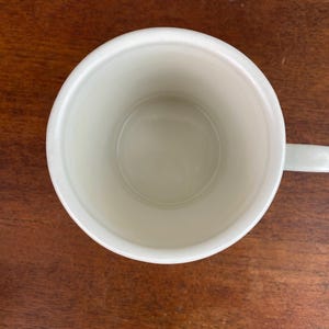 K&ouml;nnte beinhalten: Draufsicht auf eine leere, cremefarbene Keramiktasse mit Henkel. Die Tasse steht auf einer dunkelbraunen Holzoberfl&auml;che. Das Innere der Tasse ist glatt und sauber, mit leichtem Glanz.