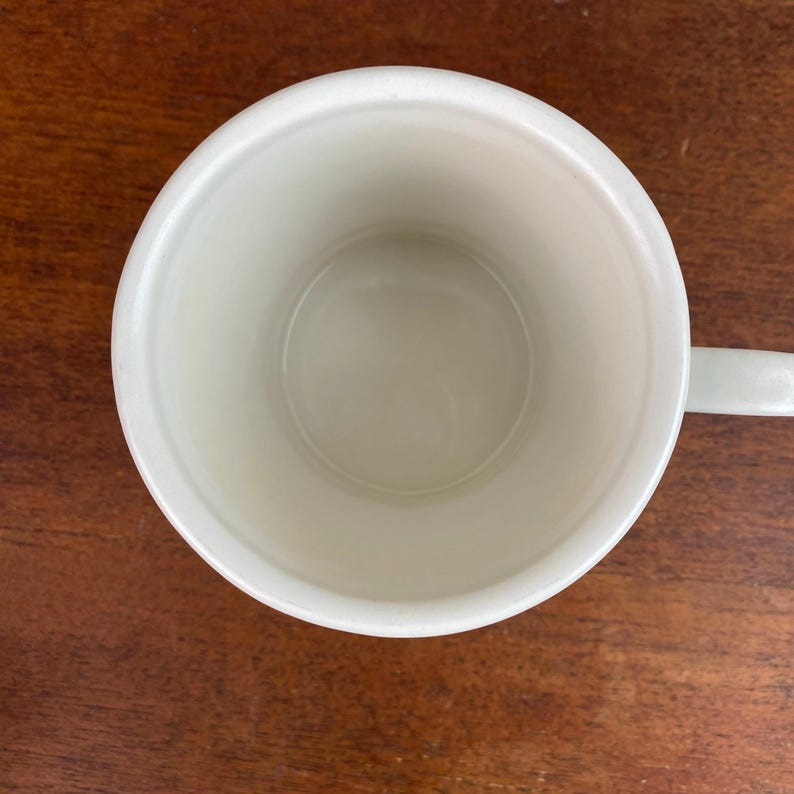 K&ouml;nnte beinhalten: Draufsicht auf eine leere, cremefarbene Keramiktasse mit Henkel. Die Tasse steht auf einer dunkelbraunen Holzoberfl&auml;che. Das Innere der Tasse ist glatt und sauber, mit leichtem Glanz.