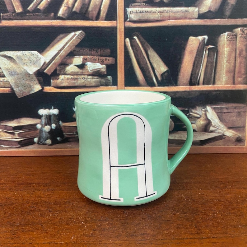 May include: A mint green ceramic mug with a white interior and a large white letter "A" on the front. The mug has a curved handle and is set against a backdrop of a bookshelf with books.