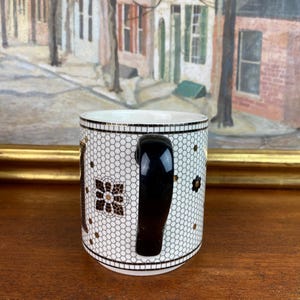 May include: White ceramic mug with a black handle. The mug has a black and gold geometric pattern.