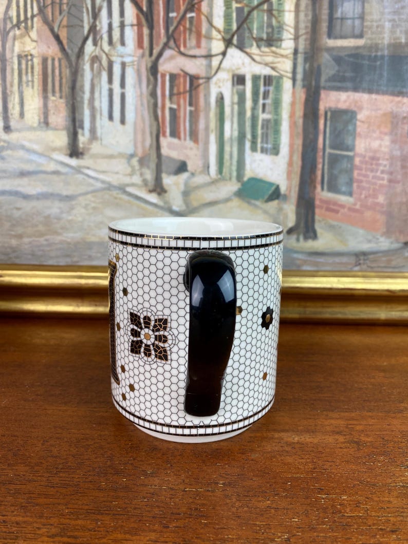 May include: White ceramic mug with a black handle. The mug has a black and gold geometric pattern.