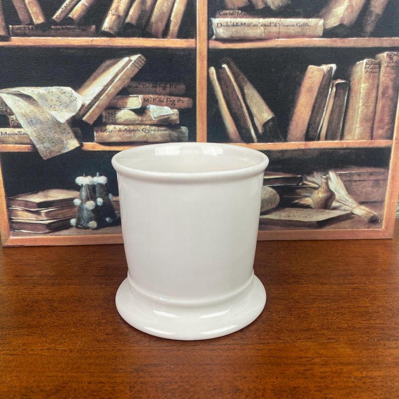 May include: A white ceramic shaving mug with a rounded base and a cylindrical body. The mug has a smooth, glossy finish and is designed for holding shaving soap or cream. The background features a painting of a bookshelf.