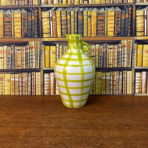 May include: A white and green chequered ceramic vase with a small handle. The vase has a rounded body and a narrow neck. The background is a bookshelf filled with books.