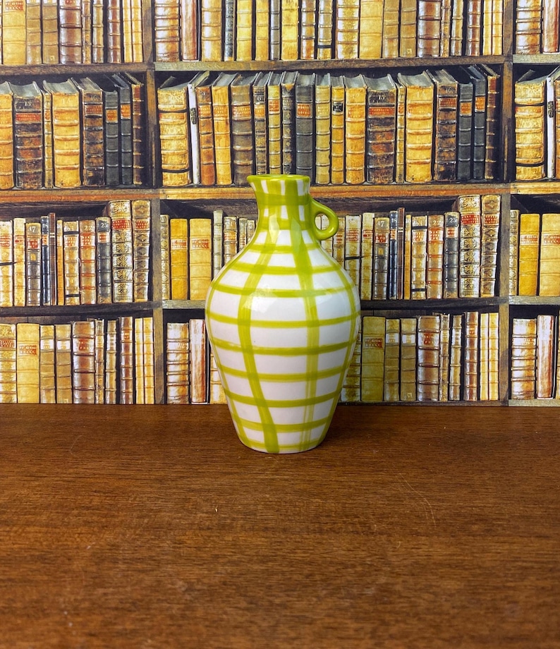 May include: A white and green chequered ceramic vase with a small handle. The vase has a rounded body and a narrow neck. The background is a bookshelf filled with books.