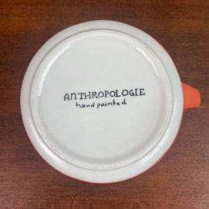 May include: A white ceramic mug with an orange exterior and handle. The bottom of the mug features the text "ANTHROPOLOGIE hand painted" in black script. The mug is set against a wooden surface.