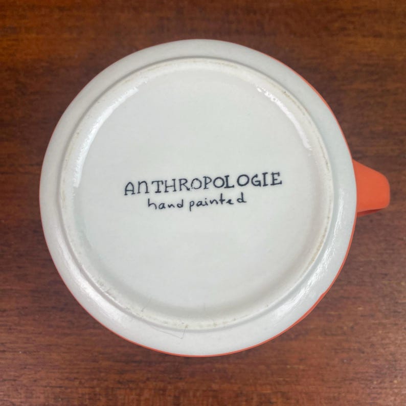 May include: A white ceramic mug with an orange exterior and handle. The bottom of the mug features the text "ANTHROPOLOGIE hand painted" in black script. The mug is set against a wooden surface.