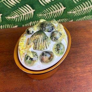 May include: An oval-shaped ceramic dish with a lid, featuring a seashell design. The dish is primarily white with a brown rim and base. The lid is adorned with various seashells in shades of green, brown, and yellow, creating a coastal theme.