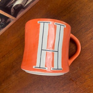 May include: Orange ceramic mug with a large white letter "H" in the center. The letter is outlined in black. The mug has a curved handle and sits on a wooden surface.