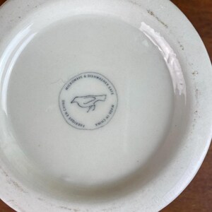 May include: A white ceramic bowl's base with a circular stamp featuring a bird illustration. The stamp reads "MICROWAVE & DISHWASHER SAFE", "FABRIQU&Eacute; EN CHINE", and "MADE IN CHINA". The bowl is on a wooden surface.
