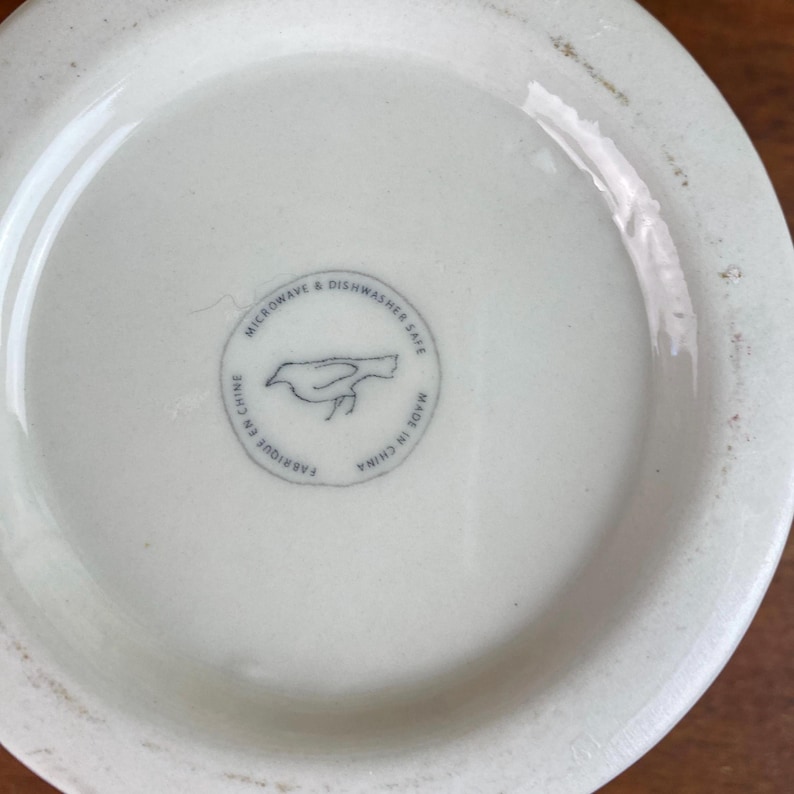 May include: A white ceramic bowl's base with a circular stamp featuring a bird illustration. The stamp reads "MICROWAVE & DISHWASHER SAFE", "FABRIQU&Eacute; EN CHINE", and "MADE IN CHINA". The bowl is on a wooden surface.