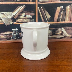 May include: A white ceramic mug with a wide base and a curved handle. The mug has a small black and white emblem on the side. The background features a bookshelf with books.