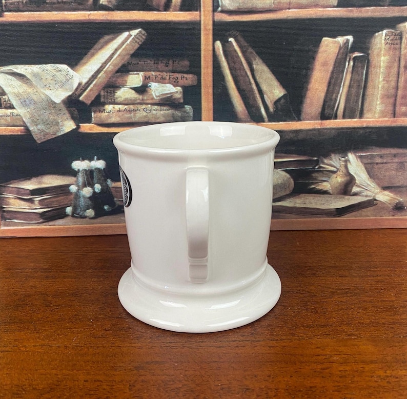 May include: A white ceramic mug with a wide base and a curved handle. The mug has a small black and white emblem on the side. The background features a bookshelf with books.