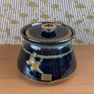 Puede incluir: Un tarro de cerámica azul oscuro con tapa, con detalles en marrón y amarillo. El tarro tiene forma redondeada y una tapa con un pomo central. El tarro tiene una etiqueta difícil de leer. El fondo es un papel pintado con flamencos dorados.