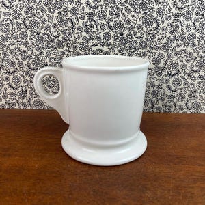 K&ouml;nnte beinhalten: Ein wei&szlig;er Keramikbecher mit Henkel und breitem Fu&szlig;. Der Becher hat ein schlichtes, klassisches Design und ist wahrscheinlich f&uuml;r Getr&auml;nke gedacht. Der Hintergrund zeigt ein Blumenmuster.