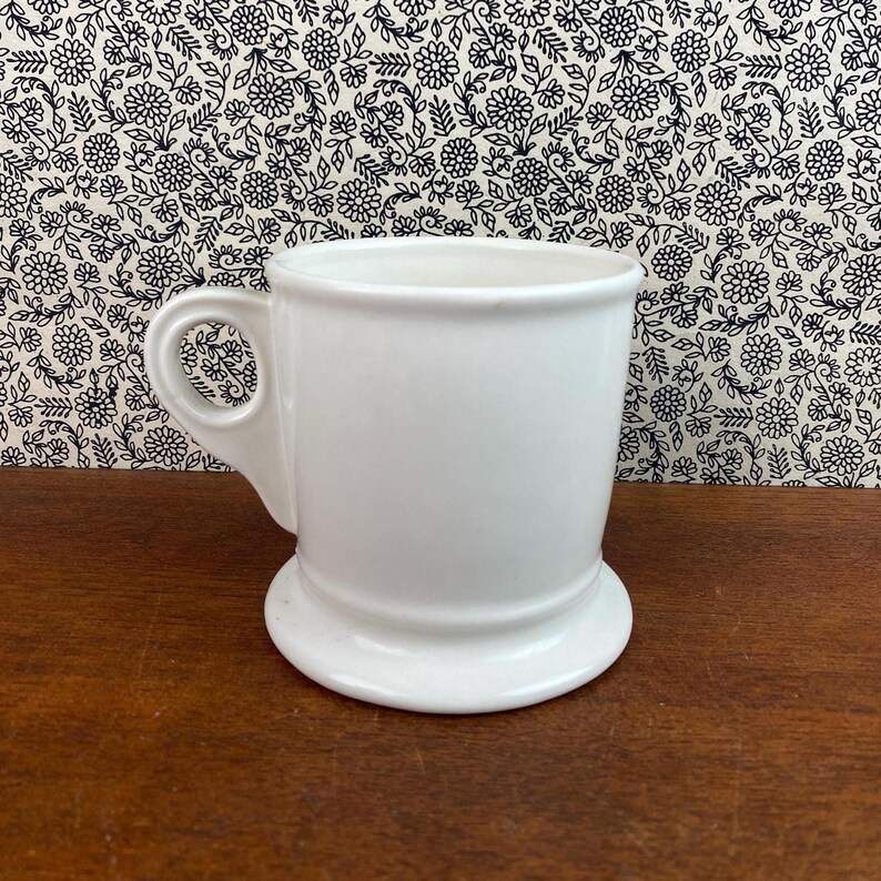 K&ouml;nnte beinhalten: Ein wei&szlig;er Keramikbecher mit Henkel und breitem Fu&szlig;. Der Becher hat ein schlichtes, klassisches Design und ist wahrscheinlich f&uuml;r Getr&auml;nke gedacht. Der Hintergrund zeigt ein Blumenmuster.