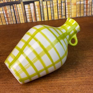 May include: A white ceramic vase with a green plaid pattern. The vase has a small handle and a flared opening. The vase is on a wooden surface.