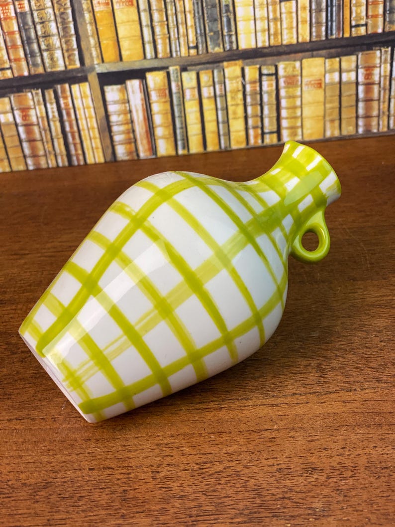 May include: A white ceramic vase with a green plaid pattern. The vase has a small handle and a flared opening. The vase is on a wooden surface.