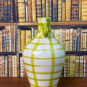 May include: A white ceramic vase with a green grid pattern. The vase has a handle and a rounded body. The background features a bookshelf with various books.