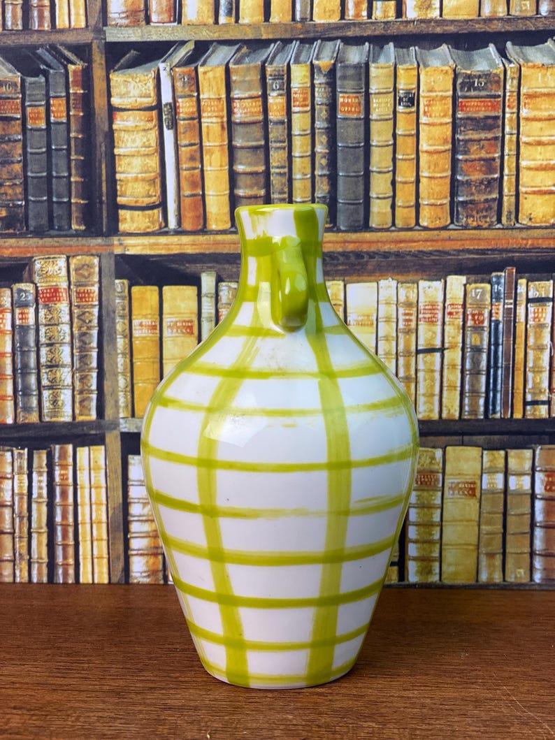 May include: A white ceramic vase with a green grid pattern. The vase has a handle and a rounded body. The background features a bookshelf with various books.