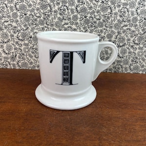 K&ouml;nnte beinhalten: Wei&szlig;e Keramiktasse mit gro&szlig;em schwarzem Buchstaben "T". Die Tasse hat einen abgerundeten Boden und einen Henkel. Der Hintergrund zeigt ein Blumenmuster. Geeignet f&uuml;r Kaffee oder Tee.