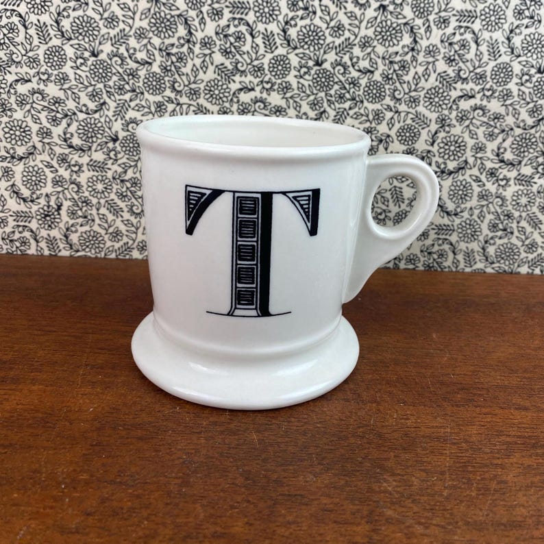K&ouml;nnte beinhalten: Wei&szlig;e Keramiktasse mit gro&szlig;em schwarzem Buchstaben "T". Die Tasse hat einen abgerundeten Boden und einen Henkel. Der Hintergrund zeigt ein Blumenmuster. Geeignet f&uuml;r Kaffee oder Tee.
