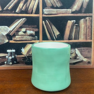 May include: A handmade, light green ceramic mug with a white interior. The mug has a rounded base and a smooth, glossy finish. The background features a vintage bookshelf with old books.