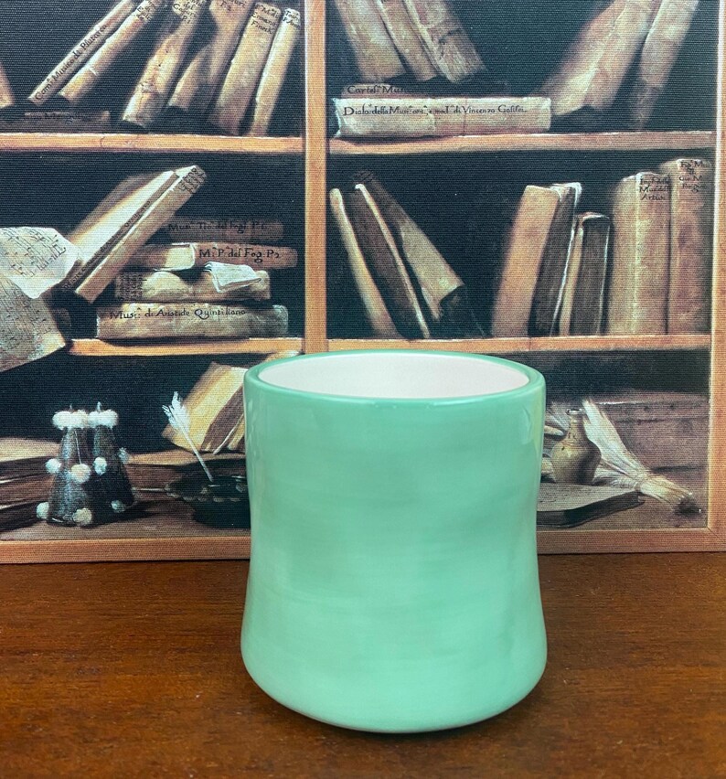 May include: A handmade, light green ceramic mug with a white interior. The mug has a rounded base and a smooth, glossy finish. The background features a vintage bookshelf with old books.