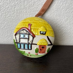 以下が含まれることがあります： わらぶき屋根の黄色い小屋、茶色のドア、緑の芝生を描いた丸いセラミックオーナメント。オーナメントには、吊り下げ用の茶色のリボンが付いています。