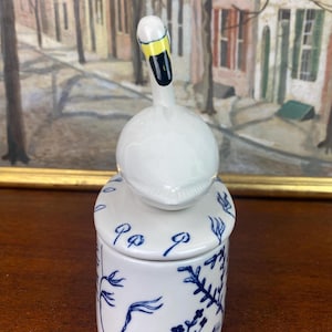 May include: A white ceramic jar with a swan-shaped lid. The swan has a yellow beak and black details. The jar is decorated with blue floral patterns and a lid with blue accents. The jar is on a wooden surface.