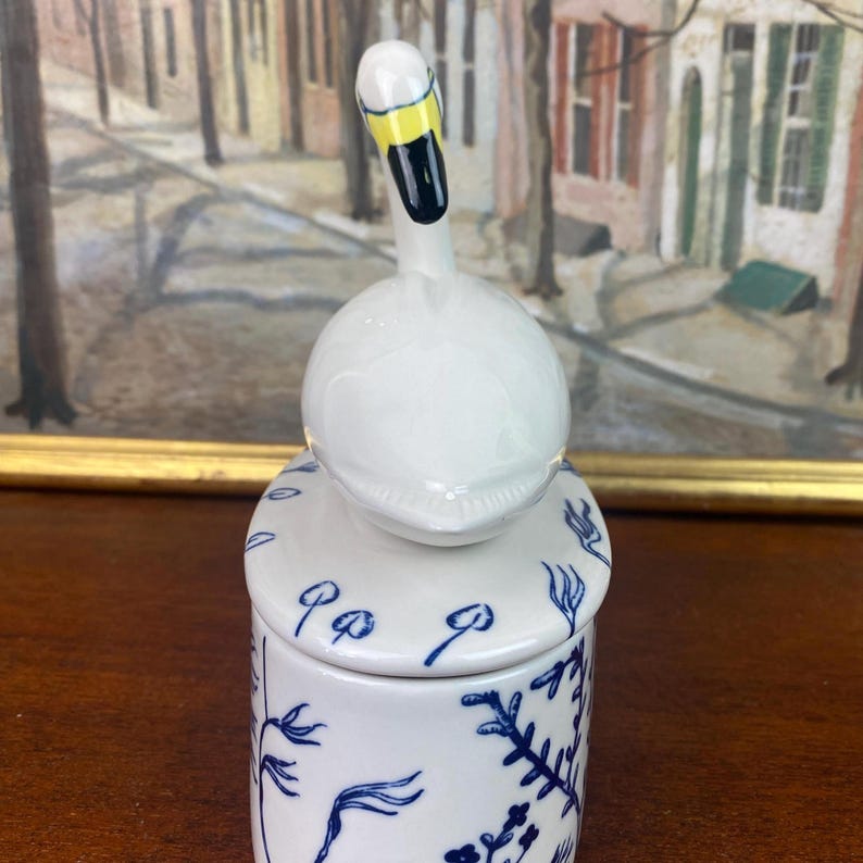 May include: A white ceramic jar with a swan-shaped lid. The swan has a yellow beak and black details. The jar is decorated with blue floral patterns and a lid with blue accents. The jar is on a wooden surface.