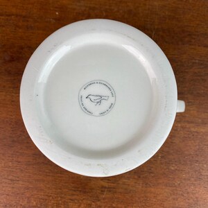 May include: White ceramic mug with a handle, seen from below. The base has a circular emblem with a bird illustration and the text "MICROWAVE & DISHWASHER SAFE", "FABRIQUE EN CHINE", and "MADE IN CHINA".
