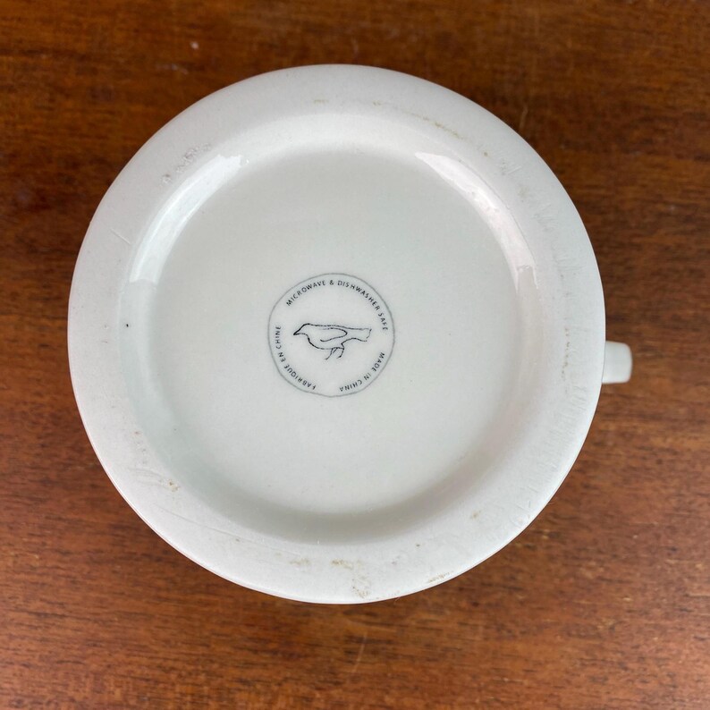 May include: White ceramic mug with a handle, seen from below. The base has a circular emblem with a bird illustration and the text "MICROWAVE & DISHWASHER SAFE", "FABRIQUE EN CHINE", and "MADE IN CHINA".