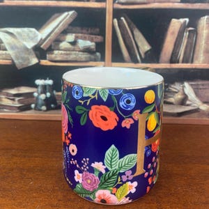 May include: A dark blue ceramic mug with a floral design featuring red, pink, and yellow flowers, green leaves, and a gold letter "F". The mug has a gold rim and a white interior.