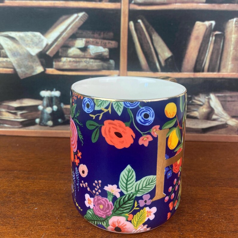 May include: A dark blue ceramic mug with a floral design featuring red, pink, and yellow flowers, green leaves, and a gold letter "F". The mug has a gold rim and a white interior.