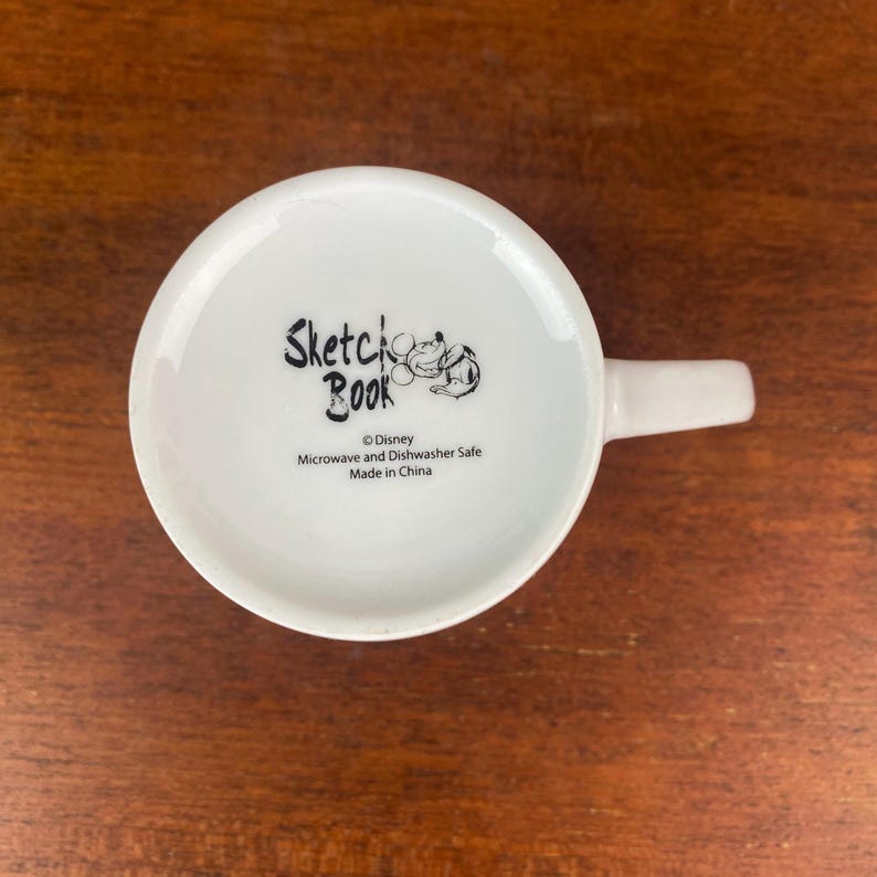 Puede incluir: Taza de cer&aacute;mica blanca con asa, con el texto "Sketchbook" y una ilustraci&oacute;n de un personaje de Disney. La parte inferior de la taza incluye el logotipo de Disney y el texto que indica que es apta para microondas y lavavajillas. Fabricado en China.