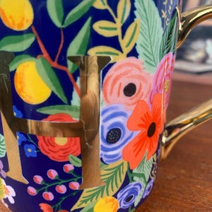 May include: A ceramic mug with a navy blue background, adorned with a floral pattern in shades of pink, orange, and blue, along with yellow lemons and green leaves. The mug features a gold handle and the gold letter "F".