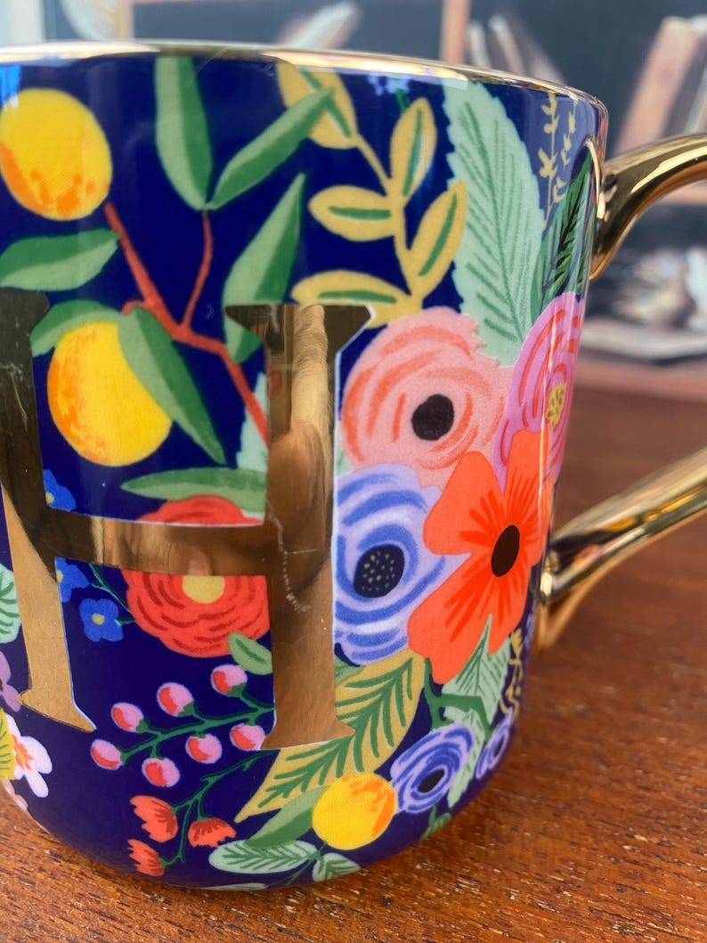 May include: A ceramic mug with a navy blue background, adorned with a floral pattern in shades of pink, orange, and blue, along with yellow lemons and green leaves. The mug features a gold handle and the gold letter "F".