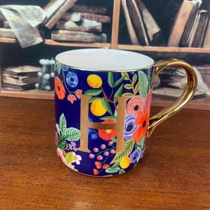 May include: A ceramic mug with a gold handle and rim, featuring a navy blue background adorned with colorful floral patterns and the gold letter "H". The mug is decorated with red, orange, and blue flowers, and yellow fruit.