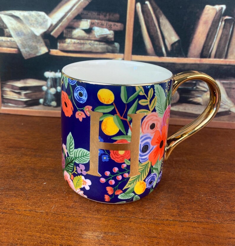 May include: A ceramic mug with a gold handle and rim, featuring a navy blue background adorned with colorful floral patterns and the gold letter "H". The mug is decorated with red, orange, and blue flowers, and yellow fruit.