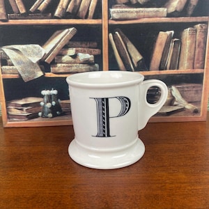 May include: White ceramic mug with a large black letter "P" on the front. The mug has a handle and a wide base. The background features a painting of a bookshelf filled with books.