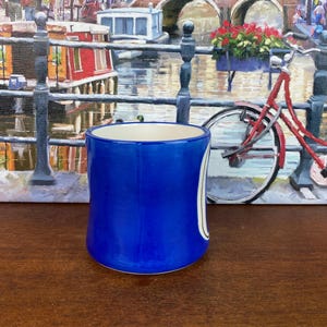 May include: A cobalt blue ceramic mug with a white interior and a white curved design. The mug is cylindrical and sits on a wooden surface. The background features a painting of a canal scene.