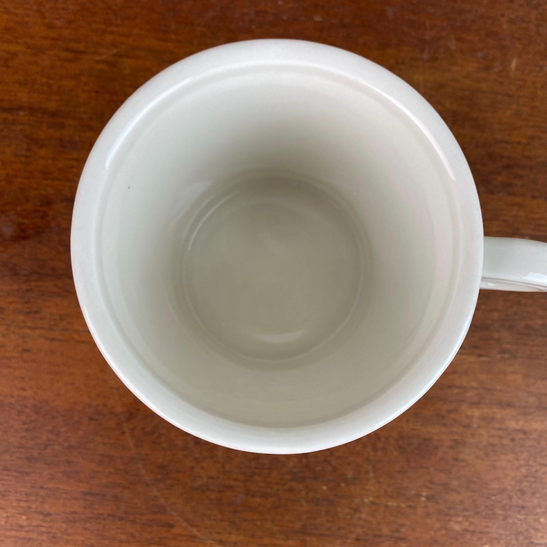 May include: A white ceramic mug with a handle, seen from above. The interior of the mug is a light cream colour, and the handle is on the right side. The mug sits on a wooden surface.