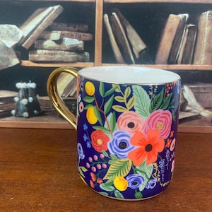 May include: A ceramic mug with a gold handle and a vibrant floral design. The mug features a dark blue background adorned with colorful flowers, leaves, and lemons. The interior is white, and the handle is a shiny gold color.