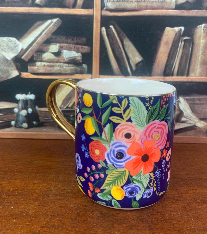 May include: A ceramic mug with a gold handle and a vibrant floral design. The mug features a dark blue background adorned with colorful flowers, leaves, and lemons. The interior is white, and the handle is a shiny gold color.